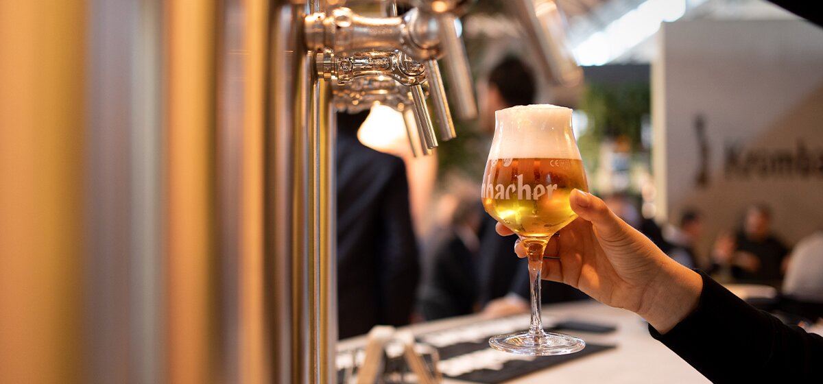 Hand holding a stemmed glass filled with golden beer topped with foam near a row of metal beer taps at a bar counter