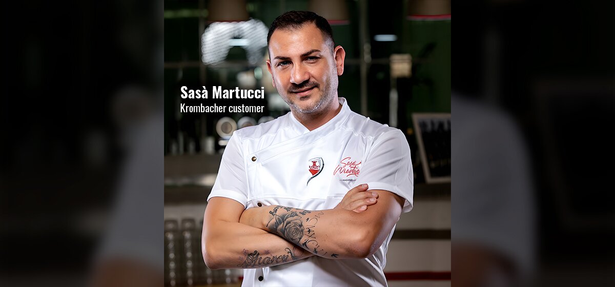 Man wearing white chef jacket with embroidered name 'Sasà Martucci' and logo, arms crossed showing floral and script tattoos on forearms, standing indoors.