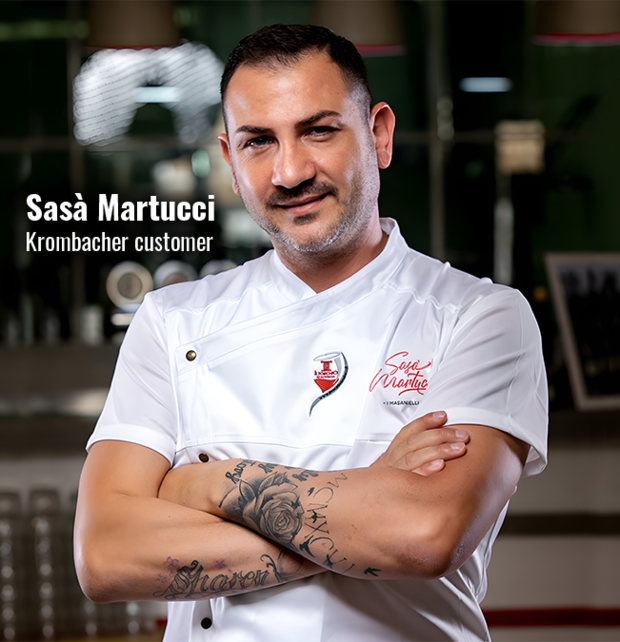 Man wearing white chef jacket with embroidered name 'Sasà Martucci' and logo, arms crossed showing floral and script tattoos on forearms, standing indoors.