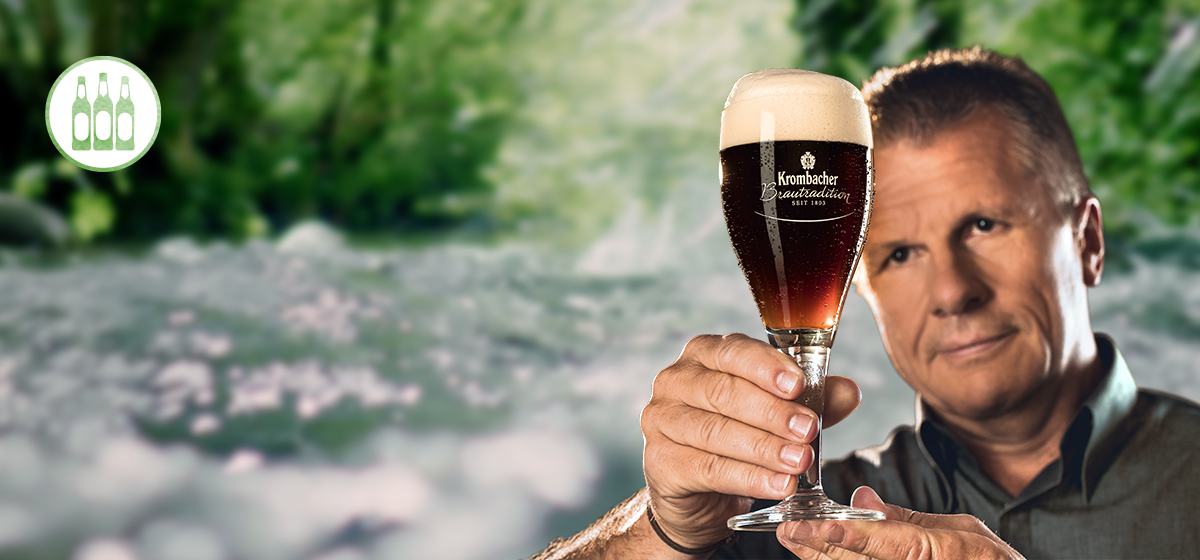 Ein Mann hält ein Glas Kombacher Bier mit Schaumkrone in der Hand. Im Hintergrund ist eine verschwommene, natürliche Landschaft zu sehen.